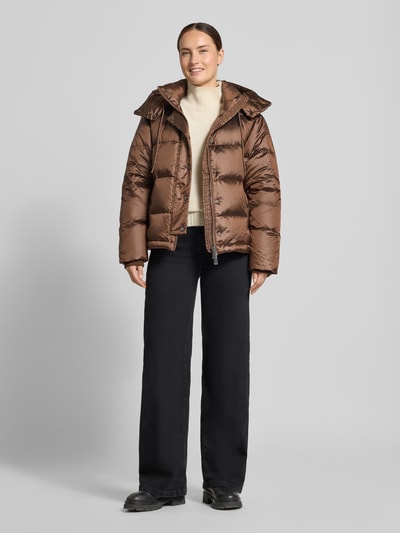 Modelka nosi kurtkę Regular Fit Jacke mit Daunen-Feder-Füllung Modell 'Jilly' od MOS MOSH, wypełnienie z puchu i pierza, z efektem pikowania, kaptur z tunelem, długość tyłu w rozmiarze S 62 cm, szerokość ramion w rozmiarze S 47 cm, szerokość w rozmiarze S 54 cm, długość rękawów w rozmiarze S 65 cm, brąz, czarne spodnie, beżowy sweter, pełny widok z przodu.