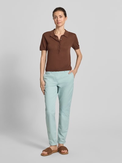 Modelka kobieca nosi Slim Fit Chino aus Baumwoll-Mix od Tom Tailor, jednolity wzór, krój slim fit, kieszenie boczne i tylne, pasek, kontrastowe szwy, zapięcie na guzik i zakryty zamek, nogawki z mankietami, bawełniany miks, widok całej sylwetki z przodu.