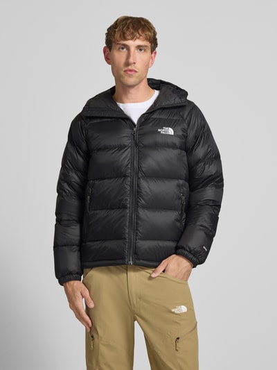 Męski model nosi kurtkę 'Jacke mit Logo-Stitching und Kapuze' od The North Face, czarny kolor, prosty krój, zamek błyskawiczny, kaptur, kieszenie zapinane na zamek błyskawiczny, wyhaftowane logo, długość tyłu w rozmiarze M 66 cm, szerokość ramion w rozmiarze M 45 cm, szerokość klatki piersiowej w rozmiarze M 55 cm, długość rękawów 70 cm, widok całej sylwetki z przodu.