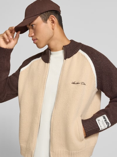 Männermodel trägt Strickjacke mit Label-Details von ANOTHER COTTON LAB in Beige und Braun, grobe Textur, Nahaufnahme Oberkörper.