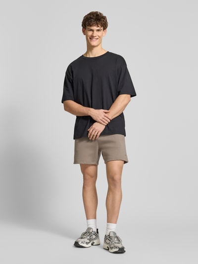 Männermodel trägt Regular Fit Shorts aus Baumwoll-Mix von Tommy Jeans in Beige mit elastischem Bund, Logo-Detail; Ganzkörperansicht von vorne.