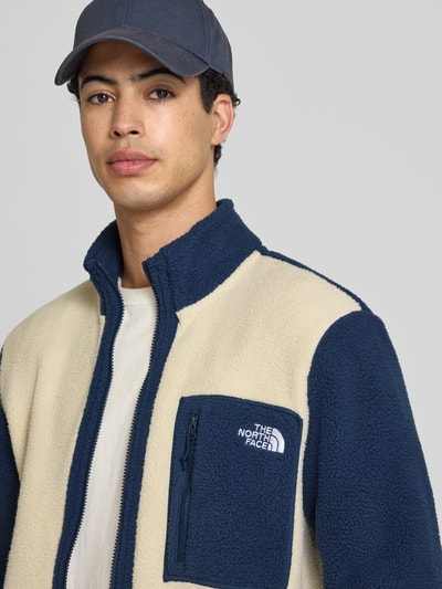 Männermodel trägt Fleecejacke mit Label-Stitching Modell 'Yumiori' von The North Face in Creme und Dunkelblau, Nahaufnahme Oberkörper, Regular Fit, Stehkragen, Reißverschluss, Reißverschlusstaschen, Rückenlänge bei Größe M 67 cm, Ärmellänge 70 cm.