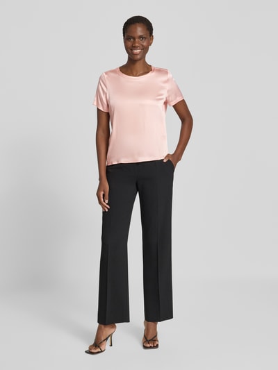 Frauenmodel trägt Blusenshirt aus Satin mit Rundhalsausschnitt von Christian Berg Woman Selection in zartem Rosa, glatte Oberfläche, kombiniert mit schwarzer Hose, Ganzkörperansicht von vorne.