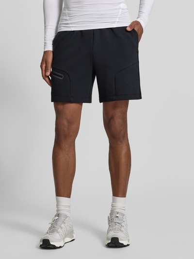 Męski model nosi szorty z Reißverschlusstasche von Under Armour, mieszanka bawełny, krój regular fit, logo, elastyczny pas, szerokość w rozmiarze M 39 cm, długość wewnętrzna nogawki 18 cm, kolor czarny, gładka tekstura, przód, ujęcie całej sylwetki.