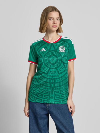 Frauenmodel trägt Mexiko Heimtrikot 2026 von adidas Sportswear, Mehrfarbig, Regular-Fit, FMF-Applikation, Kontraststreifen, Logo-Stitching, V-Ausschnitt, Ärmelform: Kurzarm, Ganzkörperansicht von vorne.