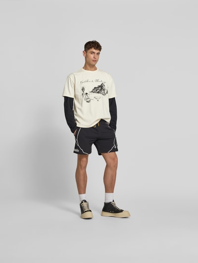 Männermodel trägt Regular Fit T-Shirt mit Motiv-Print von RHUDE in Beige, kombiniert mit Shorts, Ganzkörperansicht von vorne. Reine Baumwolle.