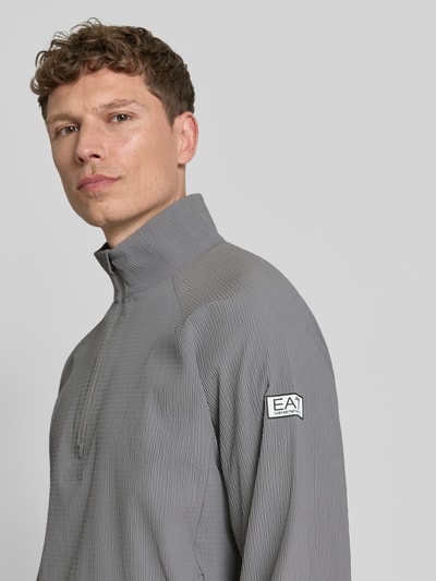 Männermodel trägt Regular Fit Sweatshirt mit Strukturmuster von EA7 Emporio Armani in Grau mit Logo-Patch, Nahaufnahme Oberkörper. Elastischer Material-Mix, Stehkragen mit Reißverschluss, strukturierte Textur.