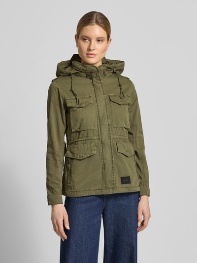 Kobiece model prezentuje kurtkę Jacke mit Pattentaschen Modell 'NORI' od khujo z bawełny, prosty krój, kieszenie z patką, funkcjonalna kaptur, stójka z zapięciem na zatrzask, ukryty zamek błyskawiczny, naszywka z logo, długość tyłu w rozmiarze S: 64 cm, szerokość ramion w rozmiarze S: 38 cm, szerokość w rozmiarze S: 48 cm, długość rękawów w rozmiarze S: 60 cm, matowa zieleń, gładka tekstura; całkowity widok z przodu