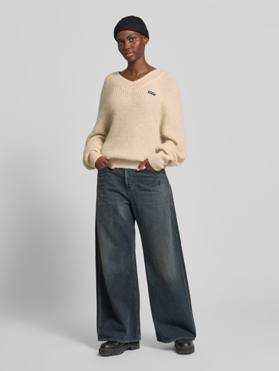 Frauenmodel trägt Oversized Pullover mit Woll-Anteil von ROTATE in Strick-Optik, V-Ausschnitt, Logo-Patch, Material-Mix, Rückenlänge 70 cm, helle Farbe, breite Form, sichtbare Textur, Ganzkörperansicht von vorne.