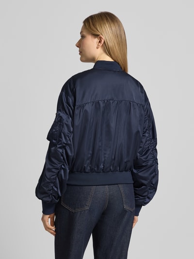 Vrouwenmodel draagt donkerblauwe Bomberjacke mit Reißverschlusstasche am Ärmel van Original Bombers, los model, opstaande kraag, ritssluiting, ritszak op de mouw, gestikt logo, zij-steekzakken met drukknoopsluiting, labelpatch, elastische boorden, ruglengte 49 cm, schouderbreedte 50 cm, borstwijdte 58 cm, mouwlengte 61 cm, gladde textuur, glanzend oppervlak, rechte vormen, ganzekörperaanzicht van achteren.