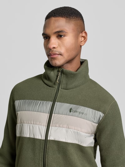 Männermodel trägt Jacke mit Stehkragen Modell 'Teca' von Cotopaxi in Olivgrün mit Label-Print, Regular Fit, Reißverschluss, Nahaufnahme Oberkörper. Rückenlänge 68 cm, Schulterbreite 43 cm, Brustweite 42 cm, Ärmellänge 63 cm, Eingrifftaschen.