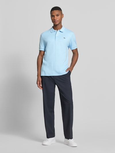 Męski model nosi Regular Fit Poloshirt aus reiner Baumwolle od Lacoste, efekt: jednolity kolor, krój regular fit, detal z logo, kołnierzyk polo, dekolt w serek, krótkie zapięcie na guziki, krótki rękaw, prążkowane zakończenie rękawów, wykończenie z rozcięciami po bokach, asymetryczny dół, materiał: czysta bawełna; widok całej sylwetki z przodu.