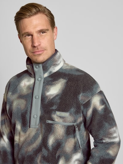 Männermodel trägt Fleecepullover mit Label-Patch Modell 'HELVETIA' von Columbia in Grau-Beige-Muster, Nahaufnahme Oberkörper. Gerader Schnitt, Stehkragen, kurze Knopfleiste, Brusttasche mit Reißverschluss, Textur weich und flauschig, Kontext: Herren Fleecepullover von Columbia.