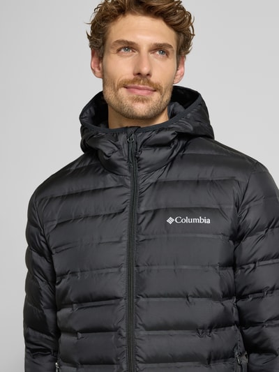 Męski model nosi czarną kurtkę pikowaną Regular Fit Model 'Lake 22™' z kapturem, nadrukiem z logo Columbia, zamek błyskawiczny, Nahaufnahme Oberkörper. Długość tyłu w rozmiarze M: 72 cm, szerokość ramion w rozmiarze M: 43 cm, szerokość klatki piersiowej w rozmiarze M: 42 cm, długość rękawów w rozmiarze M: 70 cm.