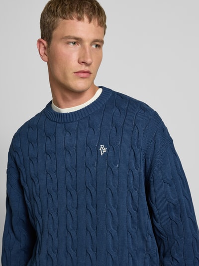 Männermodel trägt blauen Strickpullover mit Zopfmuster und Label-Stitching von REVIEW aus reiner Baumwolle, Nahaufnahme Oberkörper.