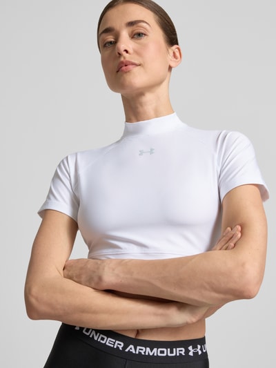 Frauenmodel trägt Cropped T-Shirt mit Logo und Stehkragen von Under Armour, weiß, Material-Mix mit Stretch-Anteil, Nahaufnahme Oberkörper. Rückenlänge bei Größe S: 30 cm.