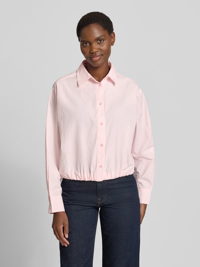 Frauenmodel trägt Regular Fit Hemdbluse aus reiner Baumwolle Modell 'Emmana' von Neo Noir, rosa, langärmlig, Hemdblusenkragen, durchgehende Knopfleiste, elastischer Saum, 1-Knopf-Manschetten, lockere Passform, Ganzkörperansicht von vorne.