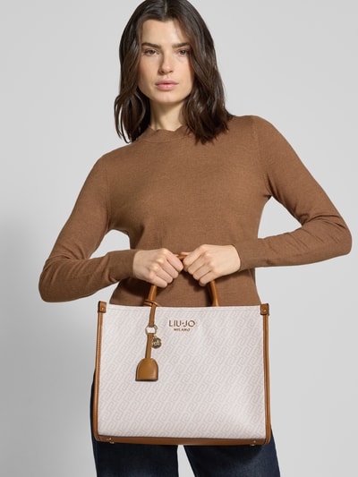 Frauenmodel trägt Handtasche mit Logo-Applikation in Leder-Optik von Liu Jo White, beiges Muster, braune Akzente, Ganzkörperansicht von vorne. Hauptfach mit Reißverschluss, Schulterriemen und abnehmbare Tragehenkel, Maße 35x27x13 cm, Länge Schulterriemen 111 cm.