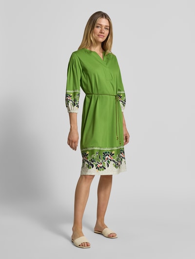 Frauenmodel trägt knielanges Kleid mit Bindegürtel von Marc Cain in Grün mit floralen Mustern, reine Baumwolle, Ganzkörperansicht von vorne.