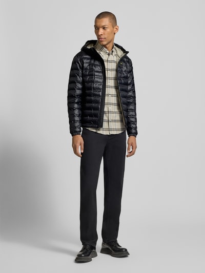 Męski model nosi czarną Kurtkę pikowaną z zamkiem błyskawicznym dwustronnym Model 'Charles' od Blauer Usa, krój regular fit, kaptur, detal z logo, długość tyłu w rozmiarze M 65 cm, szerokość ramion w rozmiarze M 47 cm, szerokość klatki piersiowej w rozmiarze M 55 cm, długość rękawów w rozmiarze M 65 cm, widok całej sylwetki z przodu.