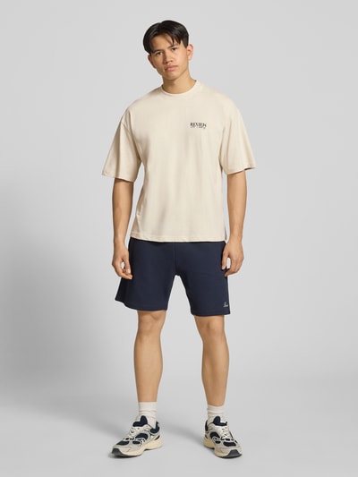 Männermodel trägt T-Shirt mit Logo-Print und Rundhalsausschnitt von REVIEW in Beige, Reine Baumwolle, Loose-Fit, Ganzkörperansicht von vorne.