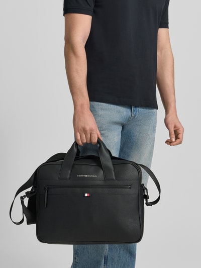 Männermodel trägt Laptoptasche in Leder-Optik von Tommy Hilfiger, schwarz, kurze Henkel, Logo, Fronttasche, Ganzkörperansicht von vorne.