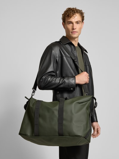Männermodel trägt Weekend Bag mit Logo-Detail Modell 'Hilo' van Rains in olijfgroen met zwarte details, Hoofdvak met ritssluiting, afneembare schouderband, Breedte 64 cm, Lengte 40 cm, Diepte 24 cm, schouderbandlengte 84 cm, Nahaufnahme Oberkörper.