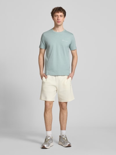 Mężczyzna nosi Shorts mit Label-Stitching mit elastischem Bund od CHAMPION w kolorze kremowym, bawełniany miks, krój regular fit, szerokość w rozmiarze M 37 cm, długość nogawki od wewnątrz w rozmiarze M 42 cm, gładka tekstura, przód, pełna sylwetka.