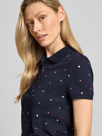 Modelka kobieca nosi Poloshirt mit Polokragen und kurzer Knopfleiste od Christian Berg Woman, bawełniany materiał ze stretchem, krój regular fit, ciemnogranatowy z kolorowymi serduszkami, długość pleców w rozmiarze S: 62 cm, zbliżenie na górną część ciała