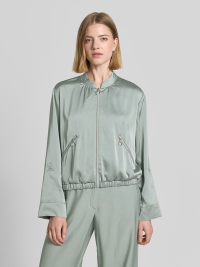 Modelka prezentuje Blouson mit regulierbarer Ärmellänge und Reißverschlusstaschen od Betty Barclay, satynowy materiał, kolor jasnozielony, krój regular fit, stójka, kieszenie z zamkiem, długość tyłu w rozmiarze 36: 53 cm, szerokość ramion w rozmiarze 36: 38 cm, szerokość w biuście w rozmiarze 36: 50 cm, długość rękawa w rozmiarze 36: 57 cm, gładka tekstura, widok z przodu, ujęcie całej sylwetki.
