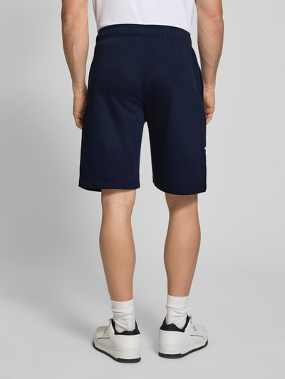 Männermodel trägt Shorts mit Logo-Print und -Stitching von CHAMPION in Dunkelblau, lockerer Schnitt, elastischer Bund, Ganzkörperansicht Rückansicht.