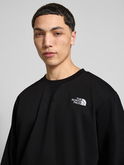 Męski model nosi Relaxed Fit Sweatshirt mit Label-Stitching Modell 'ESSENTIAL' von The North Face z bawełnianej mieszanki, czarny kolor, okrągły dekolt, elastyczne wykończenia, detal z logo, długość tyłu w rozmiarze M: 71 cm, długość rękawa: 66 cm, zbliżenie na górną część ciała.