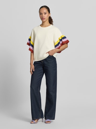 Frauenmodel trägt Boxy Fit Pullover aus Viskose-Mix Modell 'JANUARY' von Essentiel Antwerp in Creme mit bunten Streifen an den Ärmeln, weite Passform, glatte Textur, kombiniert mit weiter Jeans, Ganzkörperansicht von vorne.