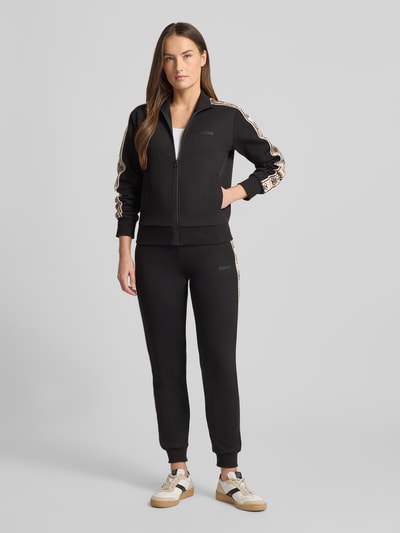 Frauenmodel trägt schwarze Regular Fit Sweatjacke mit Stehkragen Modell 'Britney' von Guess Activewear, Reißverschluss, Logo-Detail, elastische Abschlüsse, Ganzkörperansicht von vorne.