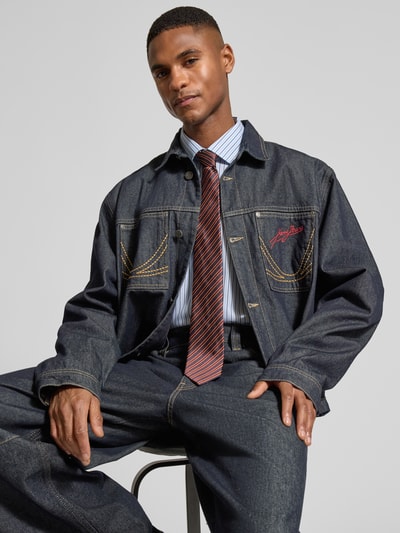 Mannelijk model draagt Relaxed Fit Jeansjacke met Ziernähten van KARL KANI in donkerblauw met contrasterende stiksels, borstzakken, label-stitching, volledig knopenpad, omgeslagen kraag, vooraanzicht bovenlichaam.