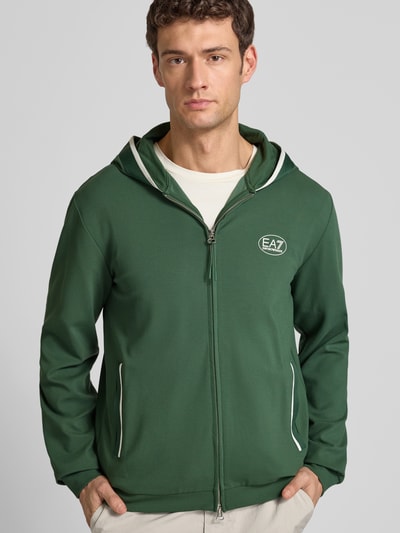 Męski model nosi zieloną Sweatjacke mit Logo und Zweiwege-Reißverschluss od EA7 Emporio Armani, kaptur, regular fit, zbliżenie na górną część ciała.