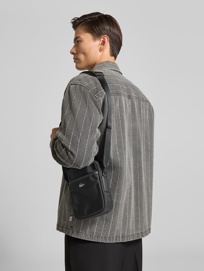 Männermodel trägt schwarze Umhängetasche mit Logo-Detail von Lacoste, kombiniert mit grauer Jacke, Ganzkörperansicht Rückansicht, Herren Umhängetasche von Lacoste.