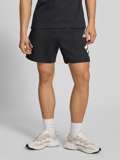 Mężczyzna model nosi Regular Fit Sport-Shorts mit Logo-Print od adidas Sportswear, kolor jednolity, elastyczny pas, kieszenie po bokach, krój regular fit, widok całej sylwetki z przodu.