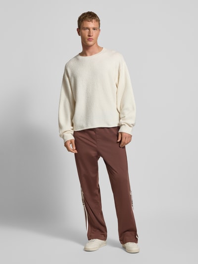 Männermodel trägt Wide Fit Jogginghose mit Logo-Stitching Modell 'Adibreak' von adidas Originals, elastischer Bund, Knopfleisten, Logo-Patch, braun, glatter Stoff, Innenbeinlänge bei Größe M 76 cm, Ganzkörperansicht von vorne.
