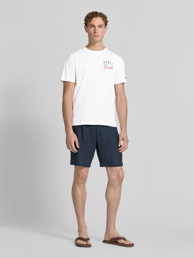 Männermodel trägt Herren T-Shirt mit Label-Prints Modell 'PORTLAND' von MC2 Saint Barth aus reiner Baumwolle, Regular-Fit, Rundhalsausschnitt, Vorder- und Rückseite mit Label-Print, Ärmel mit Label-Stitching, Farbe Weiß, kombiniert mit dunkelblauer Shorts, Ganzkörperansicht von vorne.