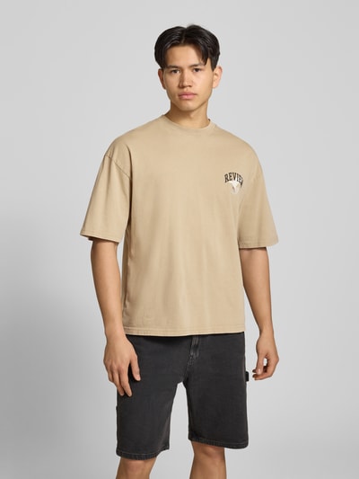 Männermodel trägt T-Shirt mit Logo- und Motiv-Print von REVIEW in Beige auf schwarzer Shorts, Reine Baumwolle, Loose-Fit, Rundhalsausschnitt, Ganzkörperansicht von vorne, Rückenlänge bei Größe M 70 cm.