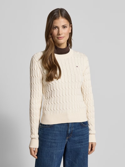 Frauenmodel trägt Slim Fit Strickpullover aus reiner Baumwolle von Tommy Hilfiger in Strickoptik mit Zopfmuster, Logo-Detail, Rundhals, Langarm, gerippten Abschlüssen, Farbe Creme, Ganzkörperansicht von vorne.