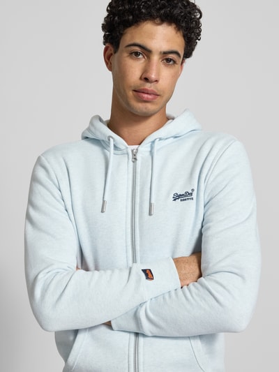 Męski model nosi Regular Fit Sweatjacke mit Logo-Stitching od Superdry, mieszanka bawełny, jasnoniebieski, zamek błyskawiczny, kieszeń kangurka, wyhaftowane logo, krój regular fit, kaptur z tunelem, elastyczne wykończenia, długość tyłu 69 cm, długość rękawa 65 cm, zdjęcie w zbliżeniu na górną część ciała.