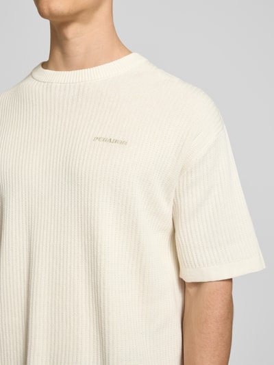 Mężczyzna nosi Strickshirt mit Strukturmuster und Label-Stitching od Pegador z bawełny, krój relaxed fit, okrągły dekolt, długość tyłu 71 cm w rozmiarze M, fakturowany wzór, wyhaftowane logo, jasny kolor, tekstura dzianiny, zbliżenie na górną część ciała.