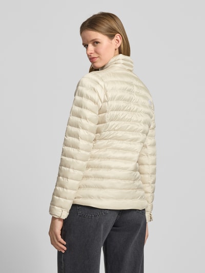 Modelka nosi kurtkę pikowaną z stójką i wyhaftowanym logo 'Steppjacke mit Stehkragen und Label-Stitching' von The North Face, krój regular fit, zamek błyskawiczny, kieszenie zapinane na zamek błyskawiczny, długość tyłu w rozmiarze S: 62 cm, szerokość ramion w rozmiarze S: 38 cm, szerokość klatki piersiowej w rozmiarze S: 46 cm, długość rękawów w rozmiarze S: 64 cm, kremowy kolor, tekstura pikowana, widok całej sylwetki z tyłu.