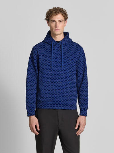 Männermodel trägt Regular Fit Hoodie aus Baumwoll-Mix von Armani Exchange in Blau mit Allover-Logo-Print und Kapuze, Ganzkörperansicht von vorne.