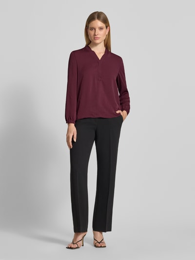 Kobieta prezentuje Regular Fit Bluse mit V-Ausschnitt od Marc Cain, bluzka o kroju regular fit, dekolt w serek, detal z logo, długość tyłu w rozmiarze 36: 65 cm, długość rękawów w rozmiarze 36: 59 cm, bordowy kolor, gładka faktura, czarne spodnie, widok całej sylwetki z przodu