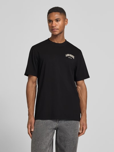 Męski model ma na sobie czarne T-Shirt mit Label-Print Modell 'Brady whop' von Les Deux z czystej bawełny, krój regular fit, okrągły dekolt, nadruk z logo, szczegół z metką, długość tyłu w rozmiarze M: 72 cm, całopostaciowe ujęcie z przodu.