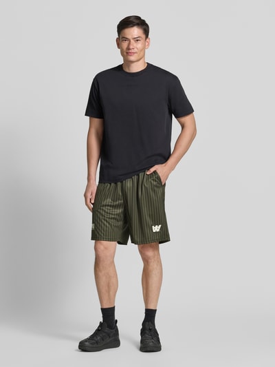 Männermodel trägt grün gestreifte Regular Fit Shorts mit Label-Print Modell 'Lama' von Wood Wood, Ganzkörperansicht von vorne, Herren Shorts, elastischer Bund, Bundweite Größe M 40 cm, Innenbeinlänge 19 cm.