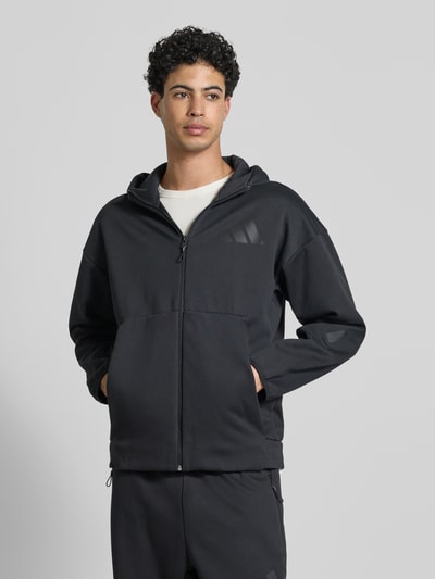 Männermodel trägt schwarze Sweatjacke mit Kapuze von adidas Sportswear, Regular Fit und Label-Print, Material-Mix, Ganzkörperansicht von vorne.